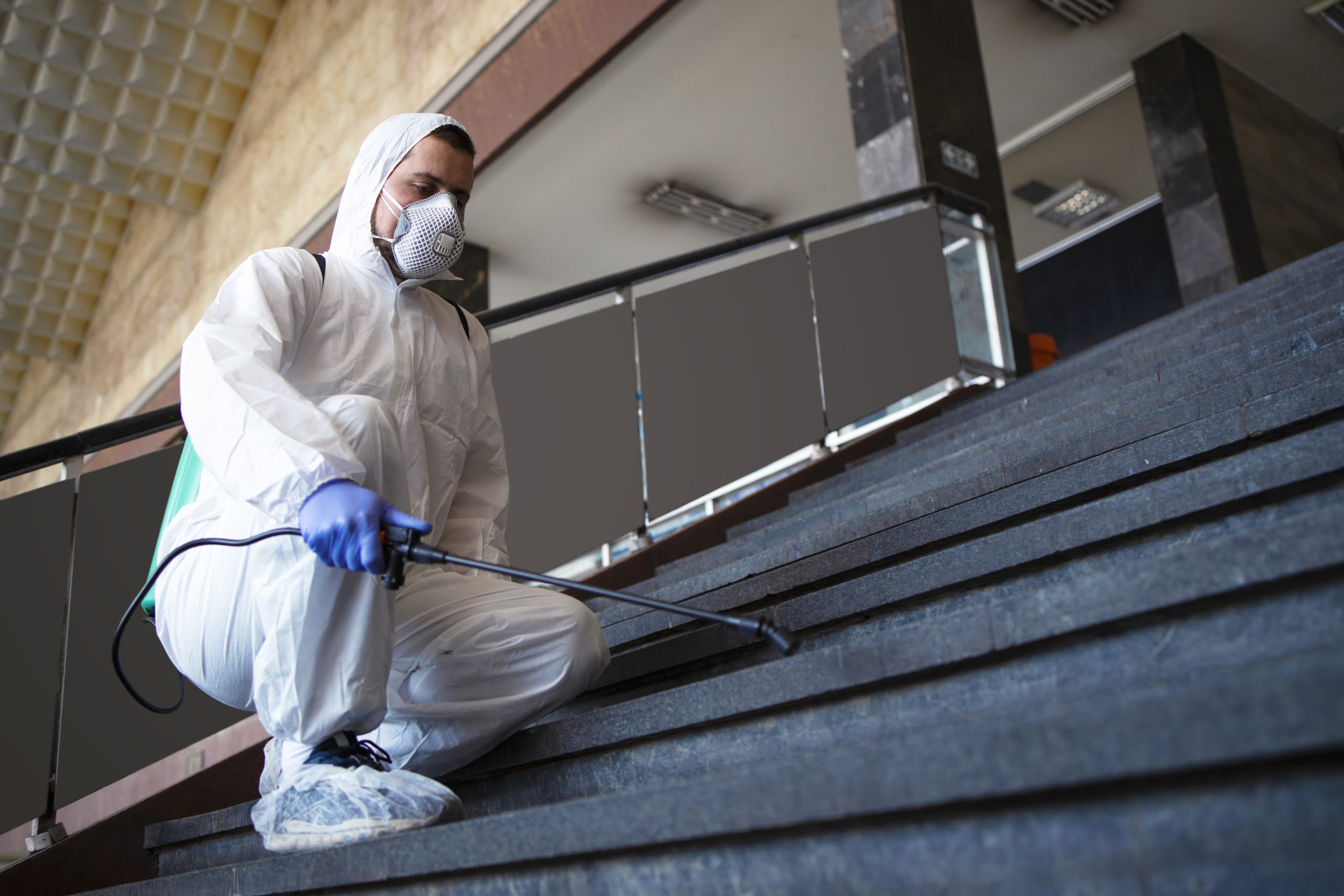 Person in white chemical protection suit disinfecting public corridors and steps to stop spreading highly contagious corona virus.